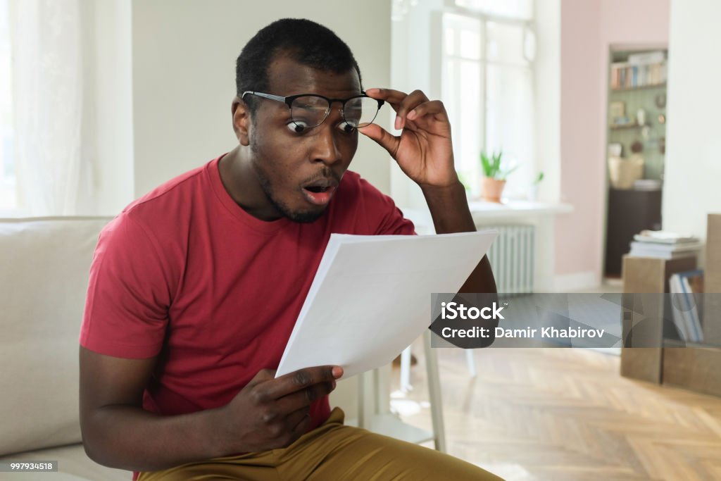 Young african american man sitting on couch in modern apartment with mouth open, holding utility bill with high rates, raised eyeglasses in wow or surprise gesture Financial Bill Stock Photo