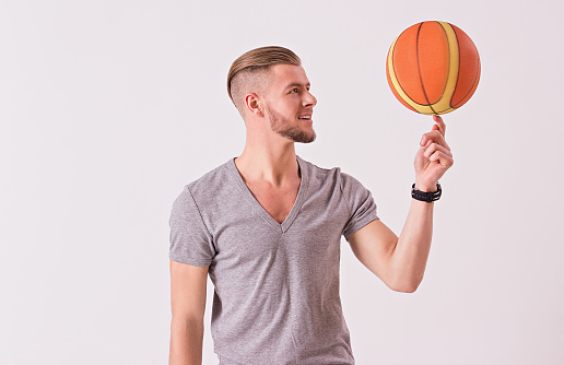 Bearded hipster in t-shirt spinning basketball ball on index finger and smiling while isolated on white background. Young sports man exercising with ball. Healthy lifestyle and sport bets concept