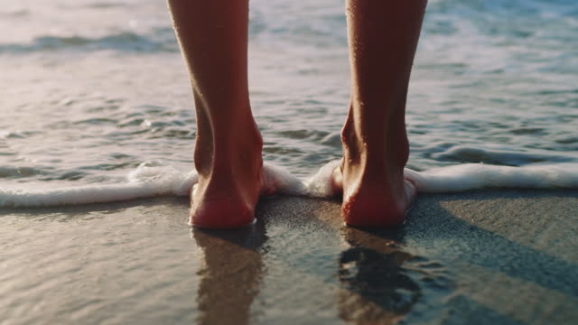 Seorang wanita muda berdiri telanjang di pantai. Kakinya mandi di ombak yang berguling di atas pasir.