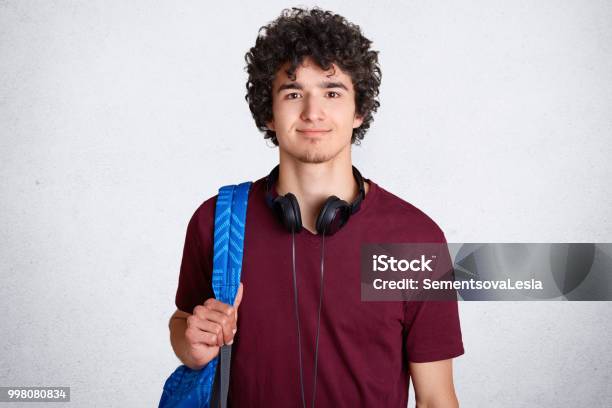 Portrait Of Delighted Hipster Male Student With Crisp Hair Wears Casual T Shirt Carries Backpack Has Headphones On Neck Ready To Go For Classes At University Isolated Over White Background Stock Photo - Download Image Now