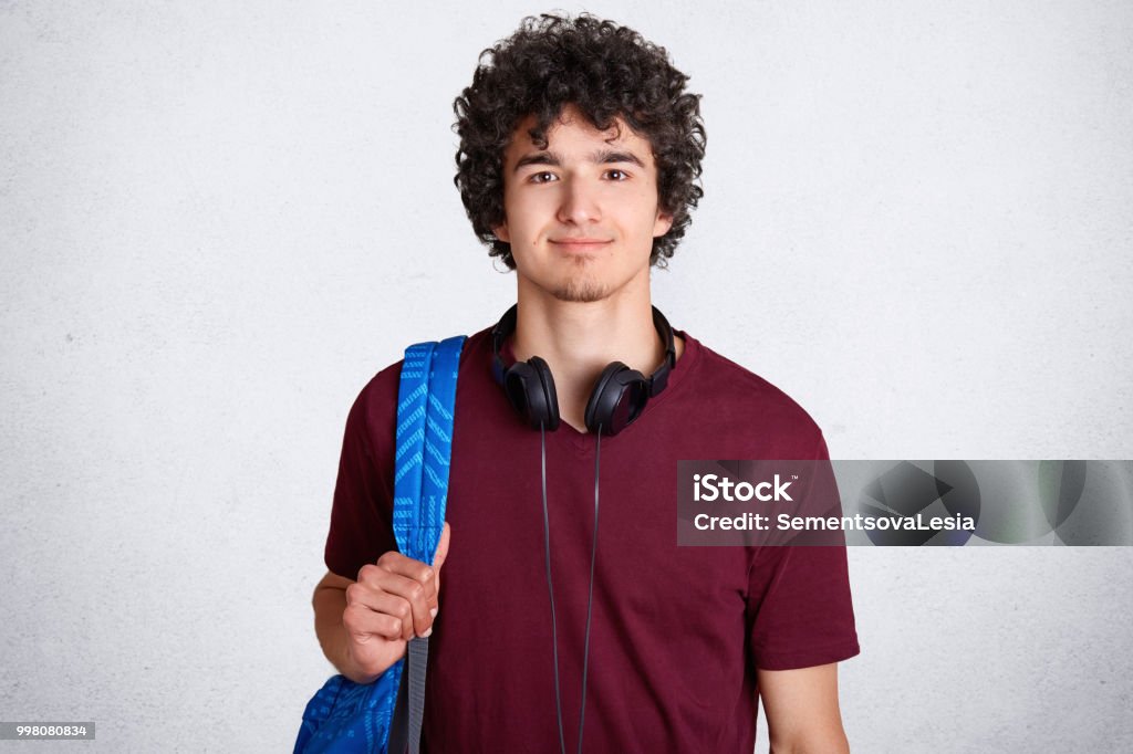 Retrato de estudante do sexo masculino hipster encantado com batata frita, cabelo, veste camisa casual t, carrega a mochila, tem fones de ouvido no pescoço, pronto para ir para as aulas na Universidade, isolado sobre fundo branco - Foto de stock de Estudante royalty-free