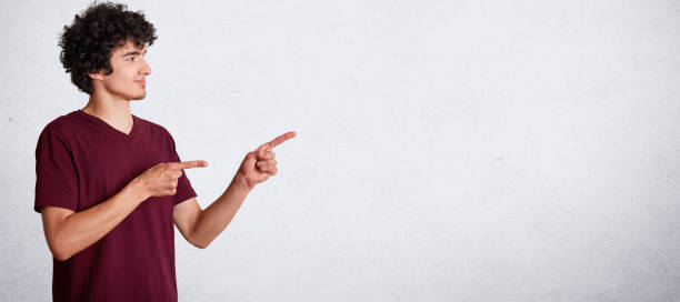 retrato horizontal de adolescente encaracolado bonito tem o cabelo escuro crocante, veste roupas casual, fica para os lados e indicar com dedos dianteiros no espaço em branco de cópia para o seu texto promocional ou propaganda - indexfinger - fotografias e filmes do acervo