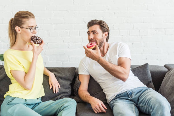 istock young couple eating donuts and sitting on sofa at home 998265446
