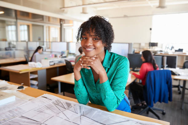 istock Black female architect leans on desk smiling in busy office 998329154