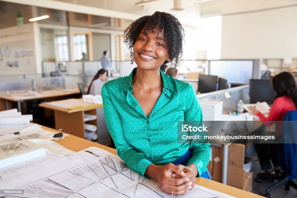Black female architect leans on desk smiling in busy office Architect Stock Photo