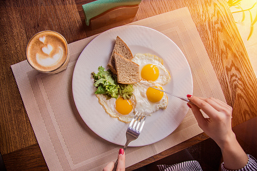 Top view of female hands holding fork and knife while having tasty breakfast at modern european cafe. Young woman eating fried eggs with toasts and morning coffee at restaurant