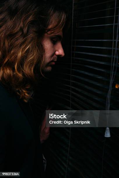 A Man With Long Hair Is Standing Near The Window Opening The Blinds With Lobes In A Black Jacket Look At The Street Spy On Peoplesunrise Stock Photo - Download Image Now - A Man With Long Hair Is Standing Near The Window Opening The Blinds With Lobes In A Black Jacket Look At The Street Spy On Peoplesunrise Stock Photo - Download Image Now -
