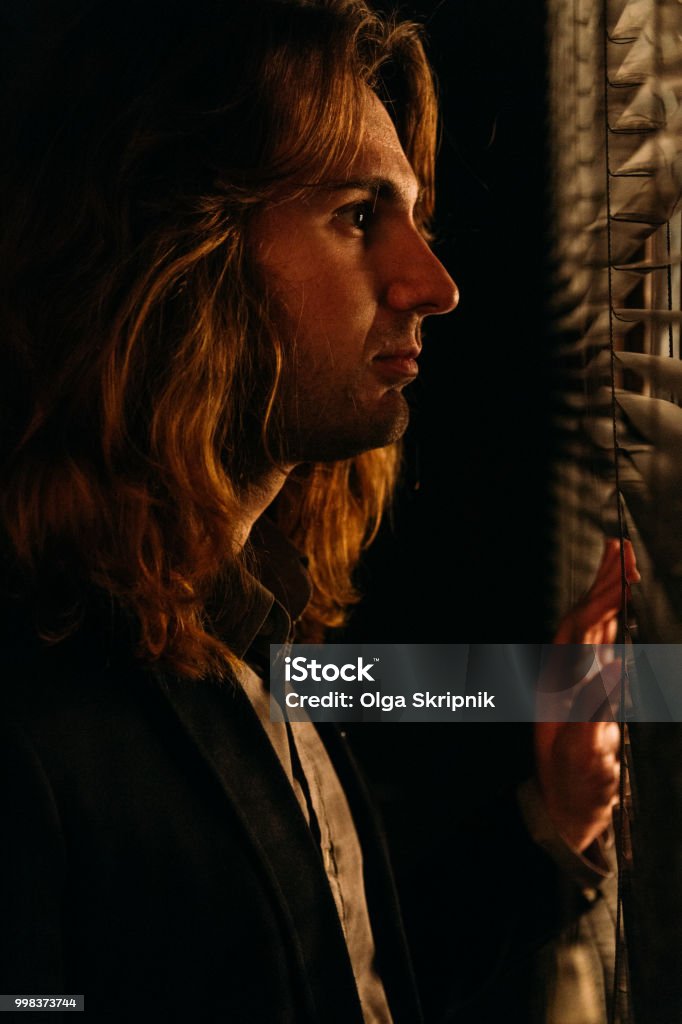 A Man With Long Hair Is Standing Near The Window Opening The Blinds With  Lobes In A Black Jacket Look At The Street Spy On Peoplesunrise Stock Photo  - Download Image Now -