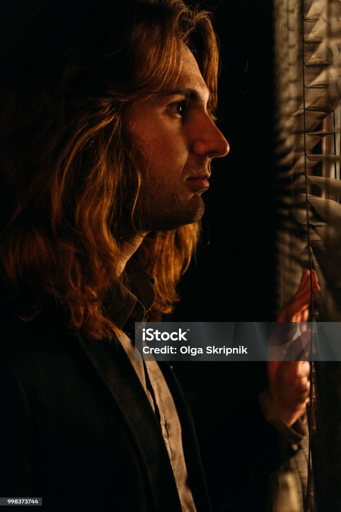 A Man With Long Hair Is Standing Near The Window Opening The Blinds With Lobes In A Black Jacket Look At The Street Spy On Peoplesunrise Stock Photo - Download Image Now - A Man With Long Hair Is Standing Near The Window Opening The Blinds With Lobes In A Black Jacket Look At The Street Spy On Peoplesunrise Stock Photo - Download Image Now -