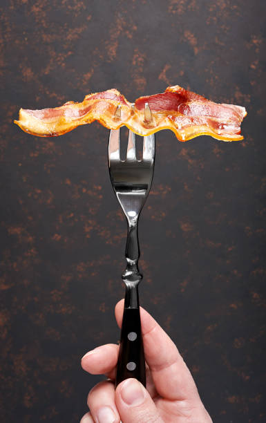 istock Fried bacon on fork in hand. Front view, isolated on black background. 998411386