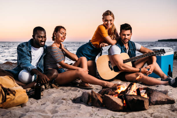 istock happy young multiethnic friends with guitar looking at bonfire while sitting together on sandy beach 998411990