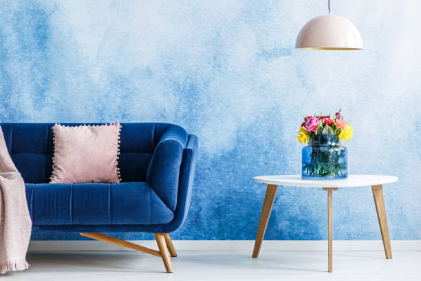 istock Comfortable plush settee with pastel cushion and a side table with fresh flowers in a vase against blue and white ombre wall in a living room interior. Lamp hanging over the table. Real photo. 998418950