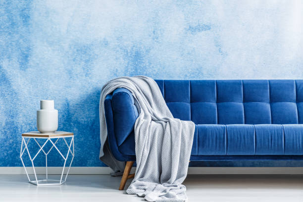 istock Navy blue sofa with gray blanket and openwork metal side table with a dish on top against watercolor, ombre wall in a modern living room interior. Real photo. 998418982