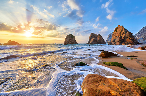 Portugal Ursa Beach at atlantic coast of Atlantic Ocean with rocks and sunset sun waves and foam at sand of coastline picturesque landscape panorama. Stones with green moss.