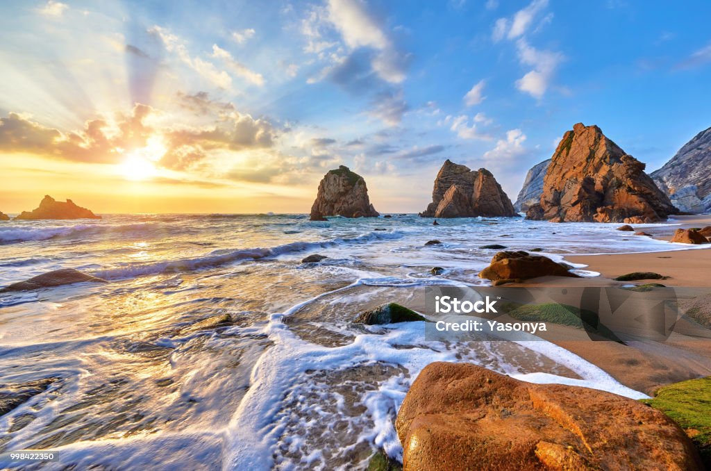 Portugal Ursa Beach sunset at Atlantic ocean Portugal Ursa Beach at atlantic coast of Atlantic Ocean with rocks and sunset sun waves and foam at sand of coastline picturesque landscape panorama. Stones with green moss. Beach Stock Photo