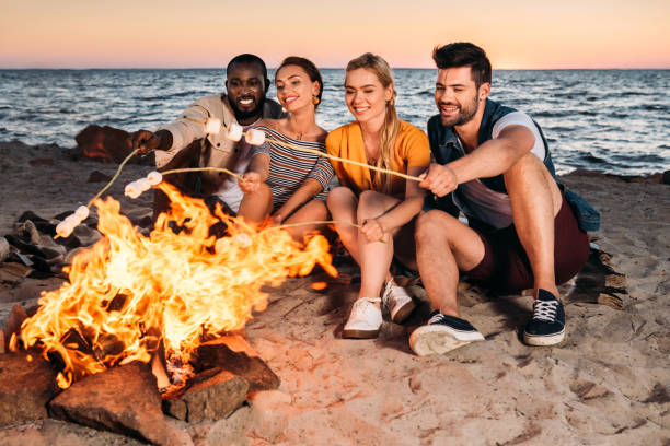 istock happy young multiethnic friends roasting marshmallows at bonfire on sandy beach at sunset 998422568