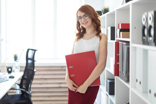 istock A beautiful young girl stands near a stack in the office and holds a folder with documents in her hands. 998457790