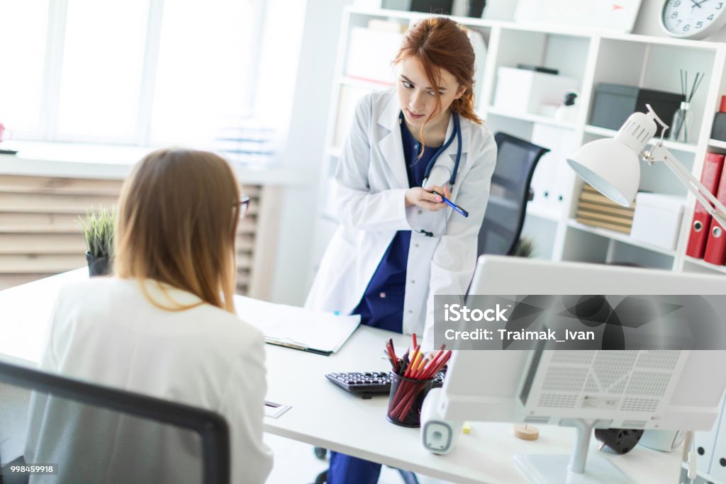A beautiful young girl in a white robe is standing near a computer desk in the office and communicating with the interlocutor. The girl makes notes in the document. A red-haired young girl with a white coat and a stethoscope on her neck. The hair is gathered in a bun. photo with depth of field Advice Stock Photo