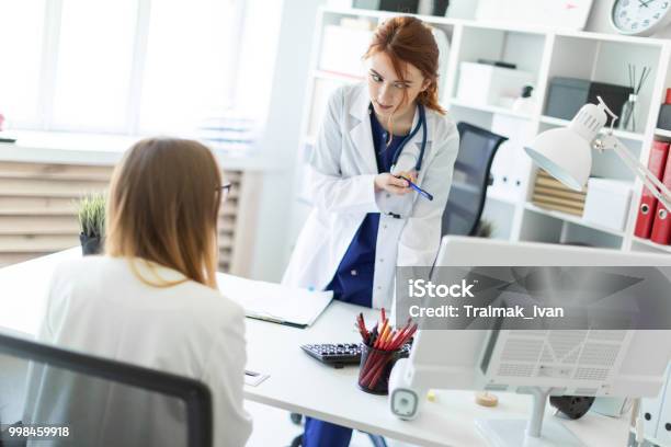 A Beautiful Young Girl In A White Robe Is Standing Near A Computer Desk In The Office And Communicating With The Interlocutor The Girl Makes Notes In The Document Stock Photo - Download Image Now
