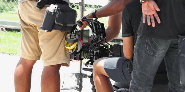 Back angle view of high definition digital video camera which recording or shooting tv commercial production on pan tilt shift professional head tripod at outdoor location and film crew team with set.