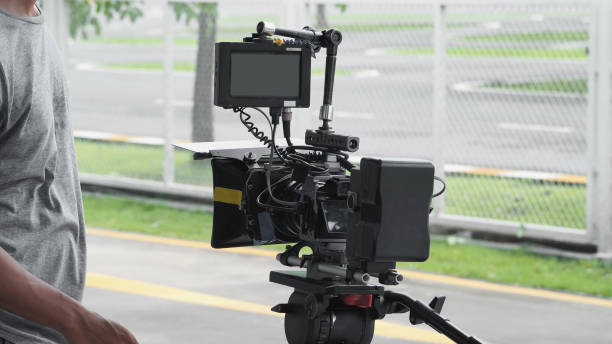 istock Back angle view of high definition digital video camera which recording or shooting tv commercial production on pan tilt shift professional head tripod at outdoor location and film crew team with set. 998488540