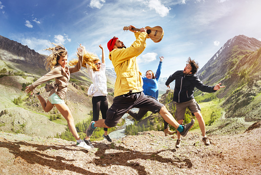 Group of happy friends with ukulele guitar is having fun and jumps against mountain during trekking. Music fun concept