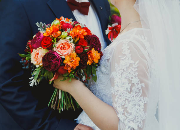 istock Plus size wedding couple is standing and hugging outside. Curvy bride is holding beautiful colorful bouquet with orange, red and pink peonies and roses. Bride and groom in summer green love story. 998509686
