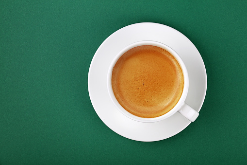 Close up one full white cup of espresso coffee on saucer on green table background, elevated top view, directly above