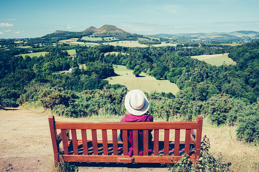 Rear view of a woman sitting on a bench at the viewpoint known as Scott's View overlooking a bend in the River Tweed towards the Eildon Hills in the Scottish Borders region of Scotland. It commemorates the novelist Sir Walter Scott.