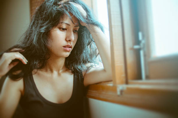 Portrait of Asian, Indian sad, serene young woman sits near window at home. She looks down, holds her head and thinks deeply with blank expression.
