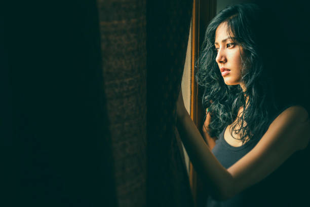 Portrait of Asian, Indian beautiful serene young woman stands near window at home. She holds the curtain, looks outside the window and thinks deeply with blank expression.