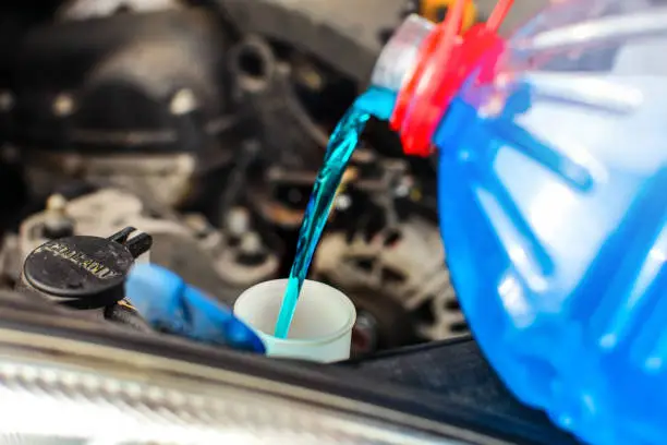 Detail on antifreeze car screen wash liquid pouring into dirty car from blue and red anti freeze water container.