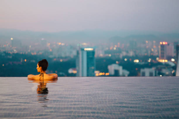 Young woman relaxing in the pool as the sun sets above Kuala Lumpur Young brunette woman relaxing in the pool, swimming on top of the building in Kuala Lumpur, Malaysia. She is having a moment to relax after work in the busy city of Kuala Lumpur, Malaysia. infinity pool stock pictures, royalty-free photos & images