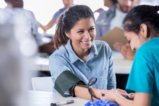 Volunteer nurse checks patient's blood pressure Young Hispanic female volunteer nurse checks a mid adult woman's blood pressure in free medical clinic. exhibition stock pictures, royalty-free photos & images