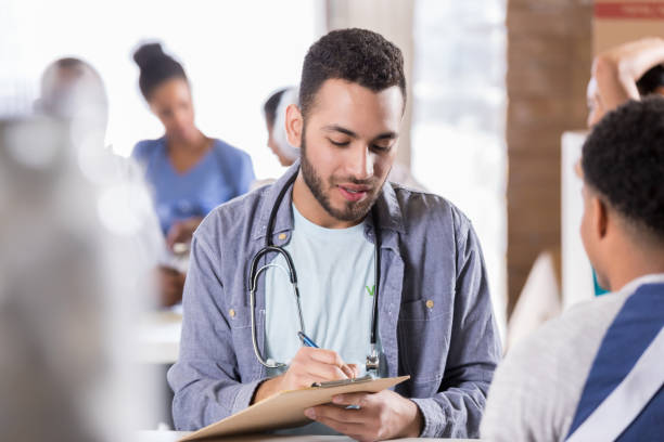 Young male doctor volunteers in free clinic Young male doctor takes notes while talking with injured African American male patient. The patient is wearing an arm sling. The doctor is volunteering in a free clinic. man with arm sling stock pictures, royalty-free photos & images