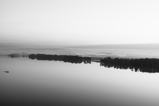 Broad river flows along diagonal shore with silhouette of forest and thick fog in grayscale. Tree drifts with flow. Minimalistic monochrome landscape of majestic nature. Morning milky atmosphere.