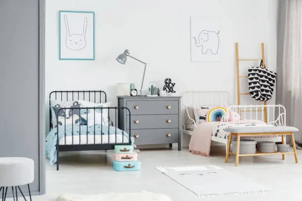 Photo of White bed for a girl and black one for a boy in a siblings bedroom interior with posters of a rabbit and an elephant on a white wall. Simple scandi design. Real photo