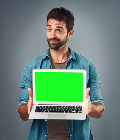 Studio shot of a handsome young man showing a laptop with a green screen against a grey background