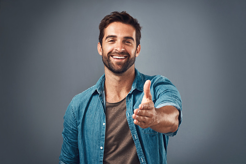 Studio portrait of a handsome young man extending his arm for a handshake against a grey background
