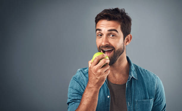 Real men eat real food Studio shot of a handsome young man eating an apple against a grey background apple bite stock pictures, royalty-free photos & images