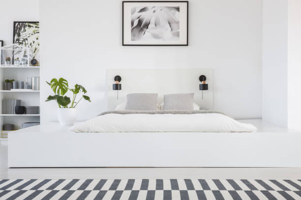 istock Real photo of a king-size platform bed standing in a white bedroom interior with a patterned rug and shelves in the bacground 998851918