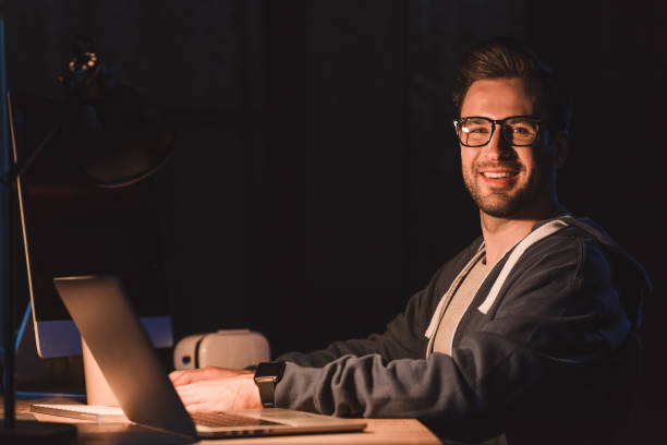 istock handsome young programmer in eyeglasses smiling at camera while working with laptop and desktop computer at night time 998855080