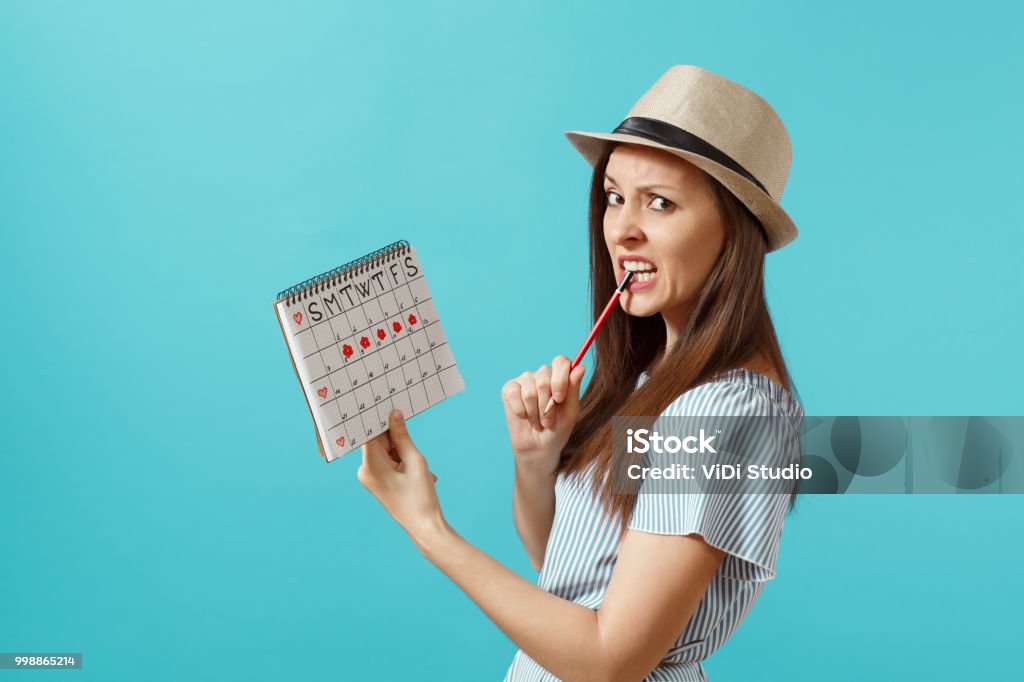 Retrato de mujer pensativa en vestido azul, sombrero con lápiz rojo, calendario femenino periodos para comprobar los días de menstruación aislado sobre fondo azul. Asistencia médica, concepto ginecológico. Copia espacio - Foto de stock de Calendario libre de derechos