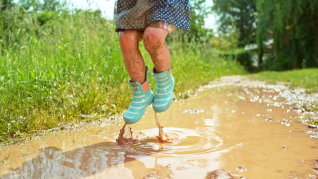 SLO MO Little girl in dotty dress and rainboots covered in mud from jumping in a muddy puddle
