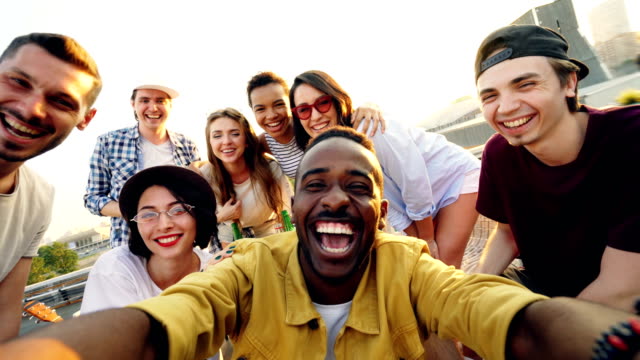Point of view shot of young people multiethnic group taking selfie and holding camera, men and women are looking at camera, smiling and posing with drinks at rooftop party.