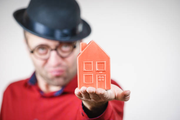 Adult man wearing funny hat and eyeglasses holding small red house model. Guy being real estate agent. Home ownership concept.