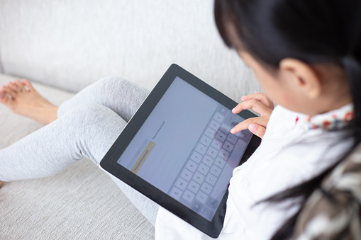 Asian Chinese little girl sitting on the sofa playing digital tablet in the living room at home