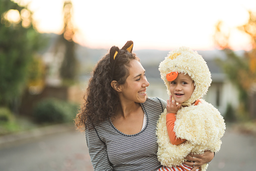 An ethnic mom holds her toddler-age boy as he smiles at the camera while they're trick or treating. He's dressed in a duck costume, she has has cat ears on.