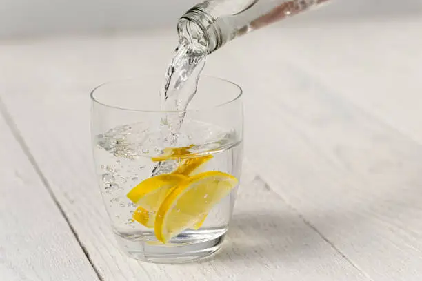 Pouring water from a glass bottle into a glass with lemon slices. White wood background.