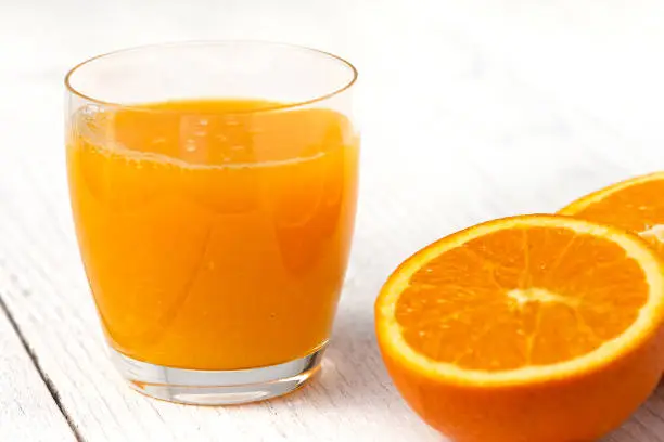 Glass of orrange juice next to halved orange. White wood background.